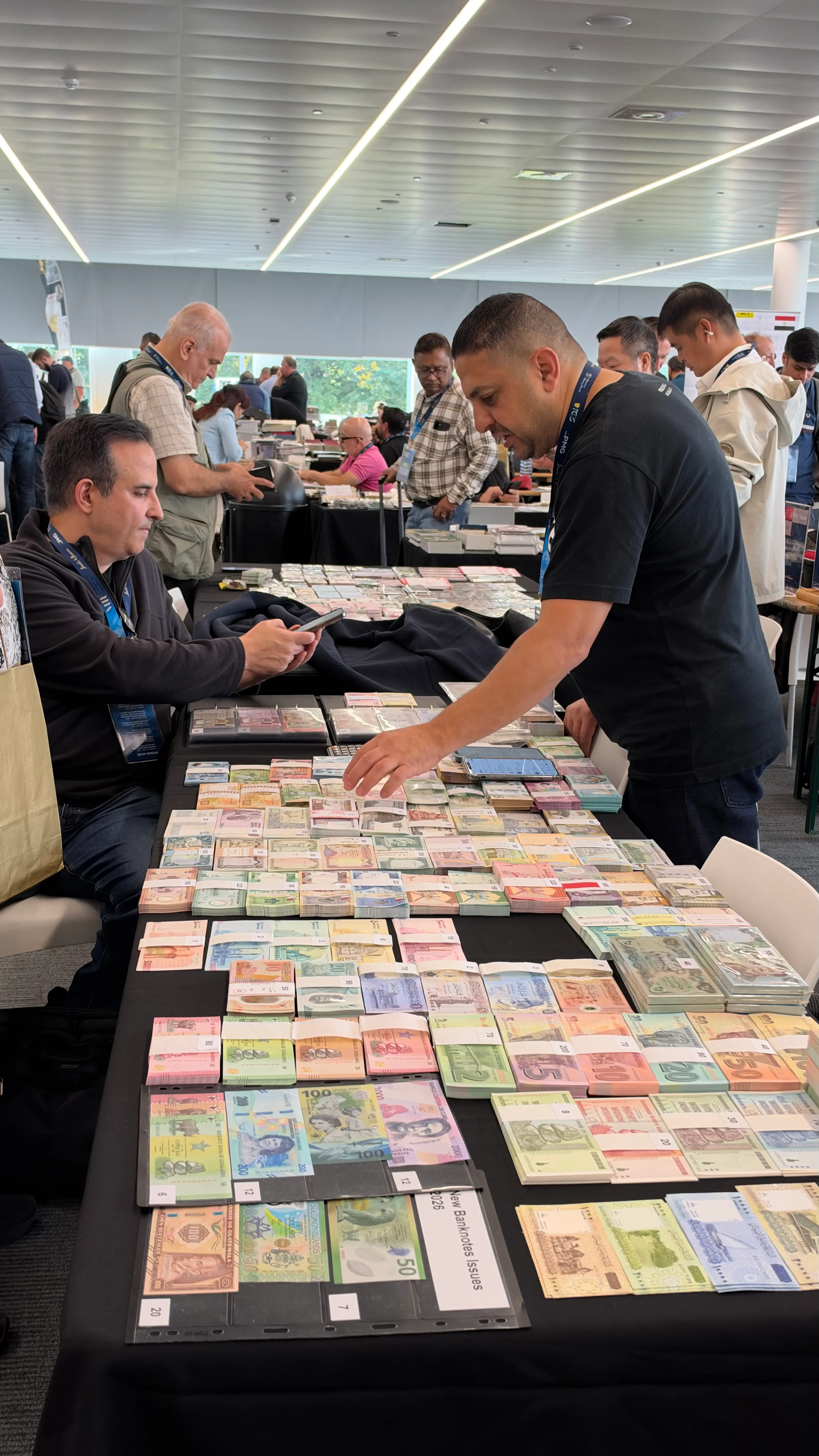 Dealer arranging bundles of new banknote issues on a table while a visitor checks his phone